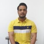 Man seated against a white wall, wearing a yellow polo with navy stripe and a tilak on his forehead.